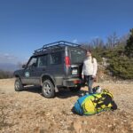 A Land Rover Discovery 4x4 vehicle parked on a rocky trail in Galicica National Park with paragliding gear and a smiling passenger.
