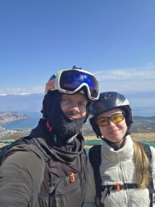 Tandem paragliding pilot and passenger wearing helmets and safety gear preparing for flight with a view of Lake Ohrid in the background.
